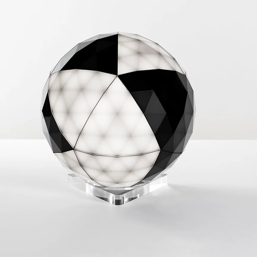 Artemide: Kugelförmiges Lichtobjekt mit abwechselnd dunklen und hellen polygonalen Flächen auf einem transparent wirkenden Sockel.