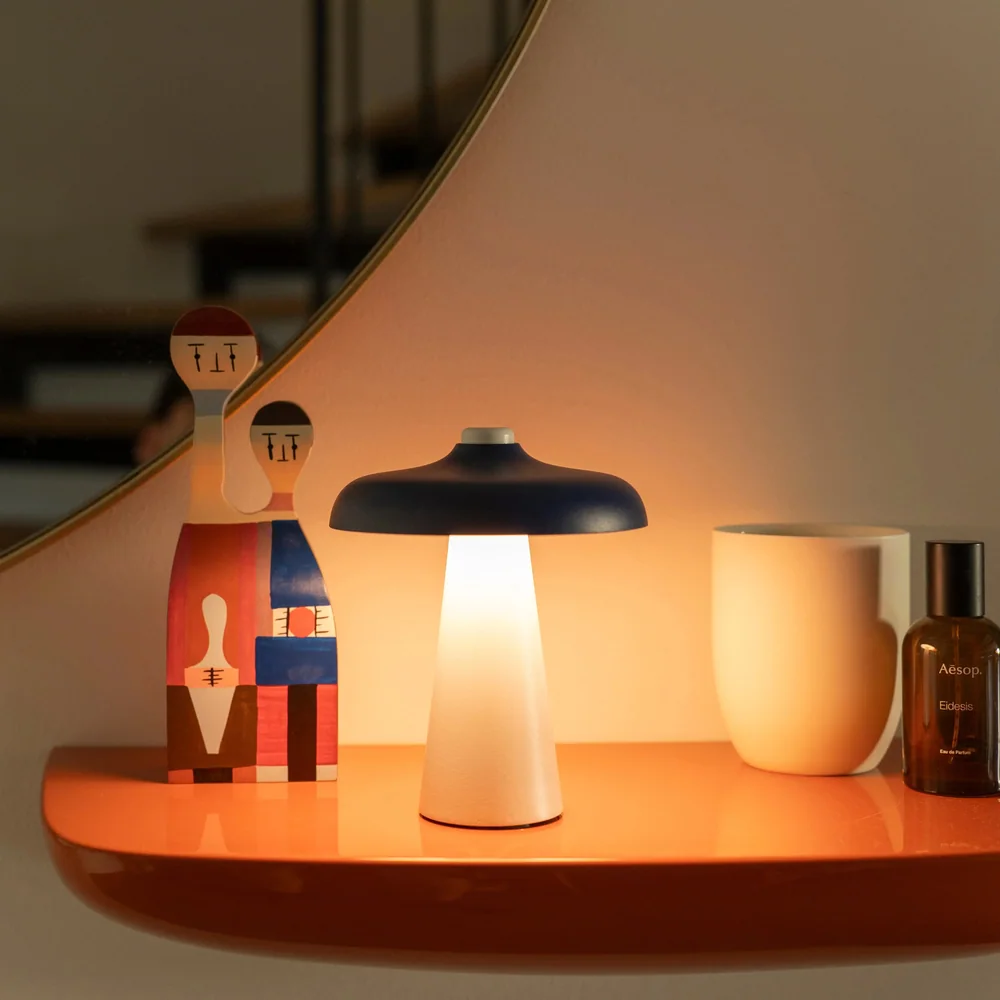 Lukuli: Kleine eingeschaltete Tischleuchte mit dunklem flachem Schirm auf einem orangefarbenen Tablett im Wohnraum.