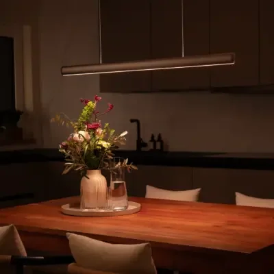 VILOOM: Bronzefarbene lineare Pendelleuchte über einem gedeckten Holztisch mit Blumen.